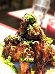 Crispy fried chicken wings piled high, glazed in sticky sauce and topped with grated cheese, sliced scallions and lemon zest on a white plate — a savory restaurant-style appetizer.
