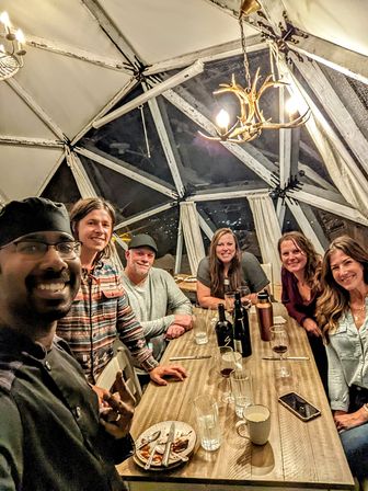 Six friends smiling in a selfie around a wooden dinner table with wine bottles and glasses inside a cozy geodesic dome at night, lit by an antler-style chandelier.