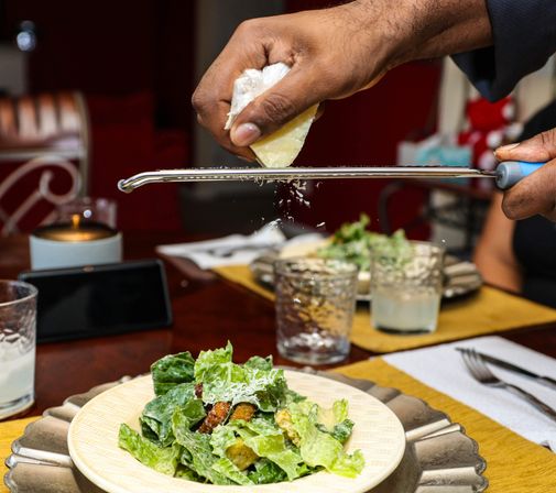 Hands grating Parmesan over a crisp Caesar salad on a cozy home dining table, cheese flakes falling onto romaine and croutons with glassware and place settings visible