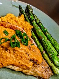 Close-up of grilled chicken breast topped with creamy roasted red pepper sauce and sliced green onions, served with mashed sweet potatoes and charred asparagus on a ceramic plate.