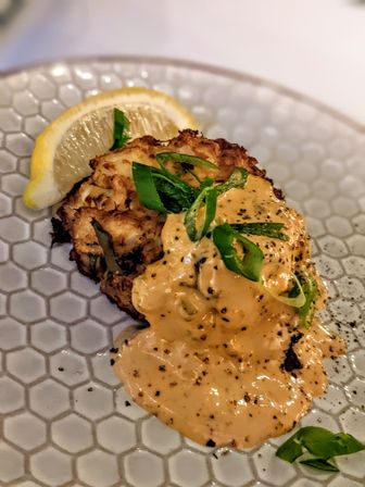 Crispy crab cake topped with creamy spicy aioli, sliced scallions and cracked pepper, served with a lemon wedge on a honeycomb-patterned plate