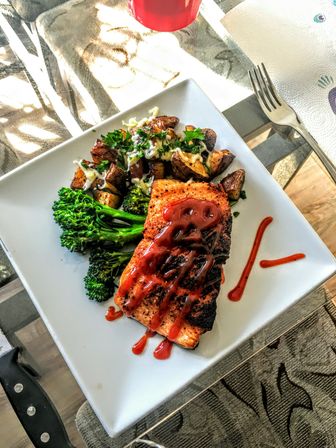 Pan-seared salmon fillet with zesty red glaze, roasted herb potatoes with melted cheese and fresh broccolini on a white square dinner plate over a glass table — colorful home-cooked healthy dinner