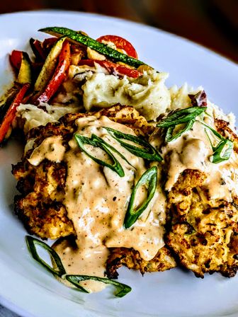 Crispy corn fritters drizzled with creamy chipotle sauce and sliced scallions, served with mashed potatoes and roasted zucchini and bell peppers on a white plate
