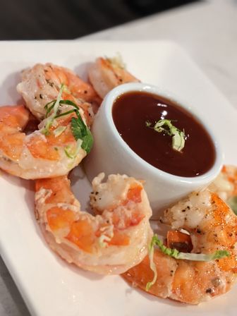 Plump grilled shrimp arranged around a small white ramekin of tangy red cocktail sauce, garnished with shredded greens on a white plate — classic seafood appetizer.