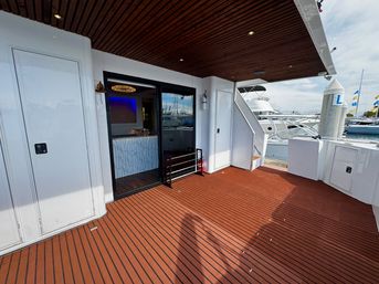 Sunny aft teak deck of a luxury yacht with wood-paneled ceiling, sliding glass salon door, white storage lockers and moored boats at a marina