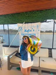 Joyful bachelorette party on a boat: woman wearing a captain hat and heart-shaped sunglasses holds a gold ring balloon in front of a 'Katie's Bachelorette' banner with water and palm trees visible outside.