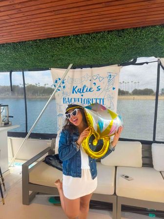 Joyful bachelorette party on a boat: woman wearing a captain hat and heart-shaped sunglasses holds a gold ring balloon in front of a 'Katie's Bachelorette' banner with water and palm trees visible outside.