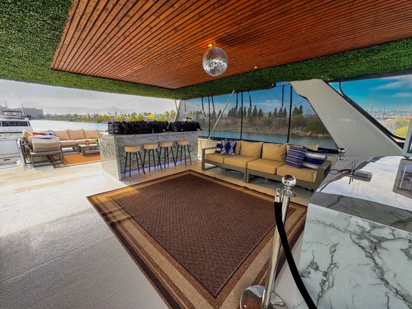 Open-air luxury yacht lounge at a marina with a marble bar and four stools, beige sectional sofas with blue pillows, disco ball under a wood-paneled ceiling with green hedge trim, woven deck rug and waterfront skyline visible through mesh screens.