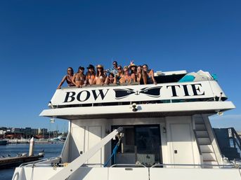 Group of friends on the upper deck of a white yacht at a sunny marina, people in swimsuits and hats leaning over the railing under a clear blue sky.