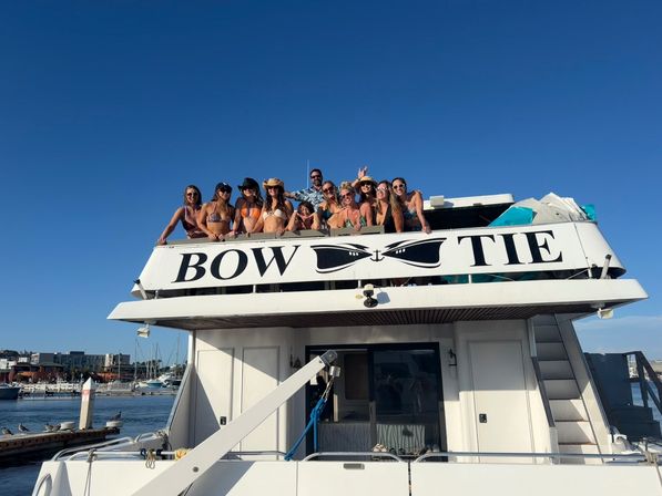 Group of friends on the upper deck of a white yacht at a sunny marina, people in swimsuits and hats leaning over the railing under a clear blue sky.