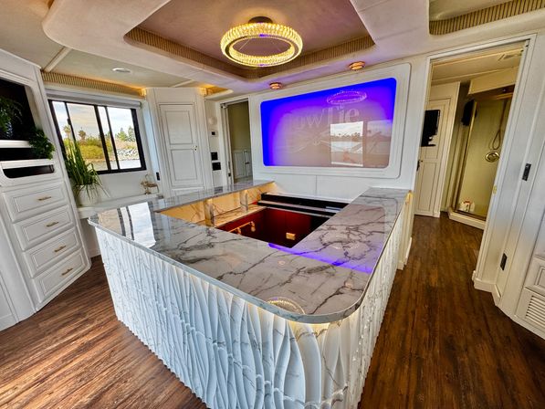 Bright luxury interior with a marble-topped island and sculpted white base, recessed bar area, purple backlit wall screen, crystal ring chandelier, wood floors, waterfront window and nearby shower