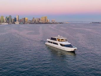 White luxury yacht cruising across calm harbor waters with the San Diego skyline and Coronado Bridge under a pastel pink-purple sunset sky