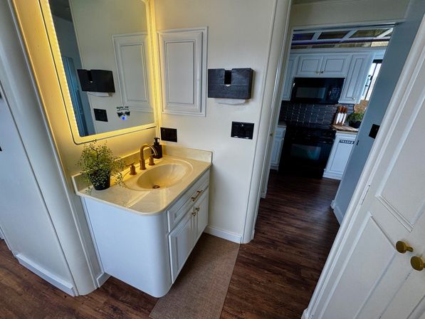 Cozy bathroom vanity with backlit mirror and warm LED glow, brass faucet, small potted plant and wall tissue holder; open doorway reveals white kitchen cabinets, black microwave and wood‑look flooring.