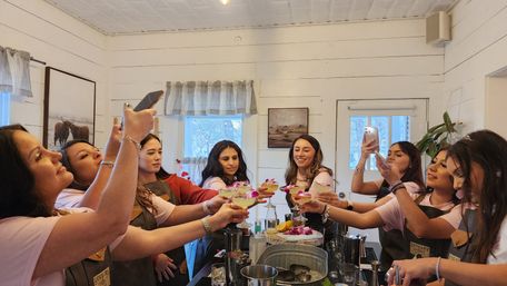 Group of women in a cozy white shiplap room toasting floral‑garnished cocktails at a hands‑on cocktail workshop, some capturing the moment on smartphones around a bar counter.