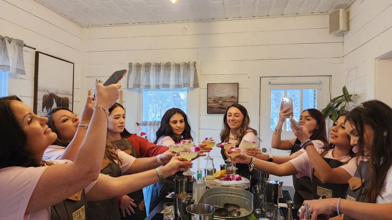 Group of women in a cozy white shiplap room toasting floral‑garnished cocktails at a hands‑on cocktail workshop, some capturing the moment on smartphones around a bar counter.