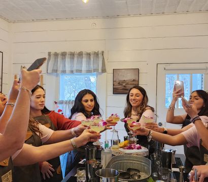 Group of women in aprons toasting floral-garnished cocktails and taking photos around a farmhouse-style kitchen island during a DIY cocktail class