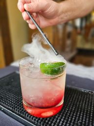 Close-up of a smoked lime cocktail in a short glass with crushed ice and red syrup at the bottom, smoke swirling as a hand uses a metal straw over a black bar mat.