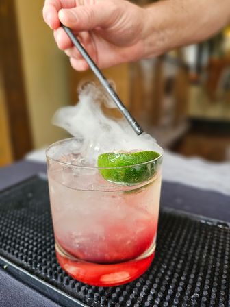 Close-up of a smoked lime cocktail in a short glass with crushed ice and red syrup at the bottom, smoke swirling as a hand uses a metal straw over a black bar mat.