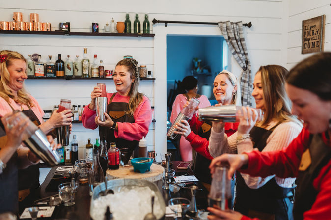 Smiling group of women in a cozy indoor cocktail-making class, wearing aprons and shaking cocktail shakers over a prep table with bottles, ice, and mixers — fun mixology workshop scene.