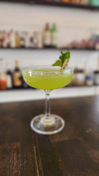 Vibrant green craft cocktail in a coupe glass garnished with a fresh herb sprig, sitting on a dark wooden bar counter with blurred bottles on shelves in the background.