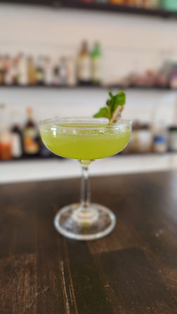 Vibrant green craft cocktail in a coupe glass garnished with a fresh herb sprig, sitting on a dark wooden bar counter with blurred bottles on shelves in the background.