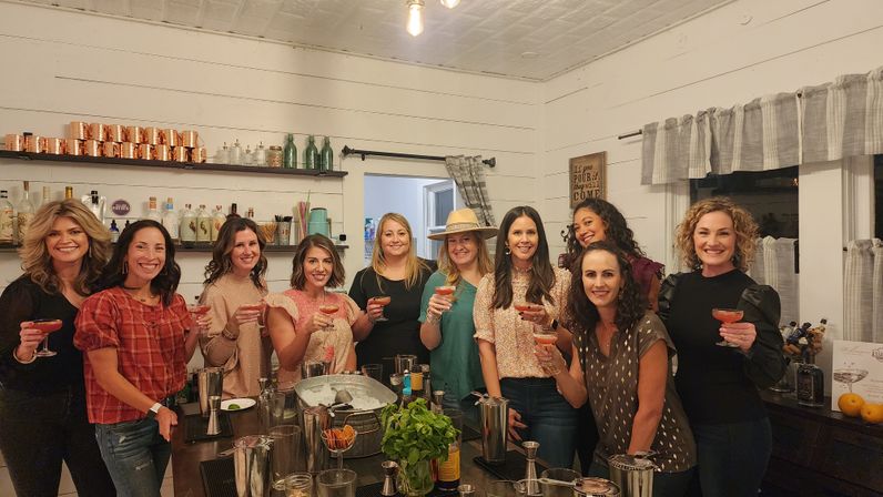 Group of women enjoying a cocktail night at a cozy farmhouse-style home bar, holding pink drinks amid copper mugs, herbs and bartending tools