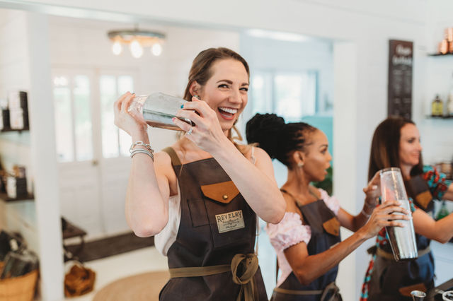 Smiling woman energetically shaking a stainless-steel cocktail shaker while wearing an apron, joining two other women in a lively mixology class in a bright modern kitchen-style bar