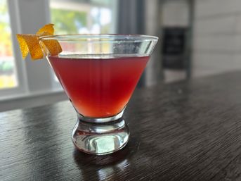 Vivid red cocktail in a short martini glass topped with an orange peel twist, resting on a dark wood bar top with a sunlit window blurred in the background