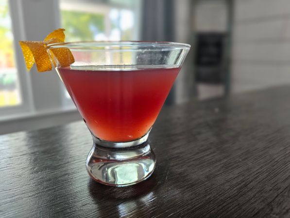 Vivid red cocktail in a short martini glass topped with an orange peel twist, resting on a dark wood bar top with a sunlit window blurred in the background