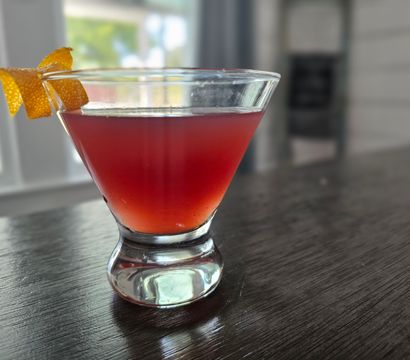 Vibrant red cocktail in a stemless martini glass garnished with an orange twist on a dark wood countertop by a sunlit window.