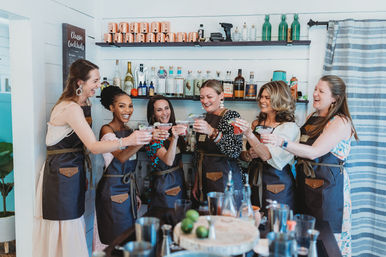 Seven women in aprons laughing and toasting colorful cocktails at an indoor mixology workshop, bar counter with bottles, copper mugs and limes.