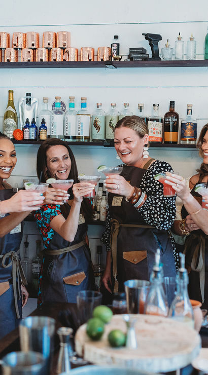 Seven women in aprons laughing and toasting colorful cocktails at an indoor mixology workshop, bar counter with bottles, copper mugs and limes.