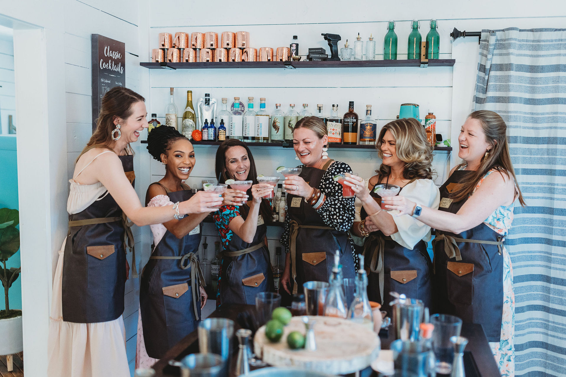 Seven women in aprons toast with colorful cocktails behind a bright bar counter during a lively mixology class, with shelves of bottles and copper mugs in the background.