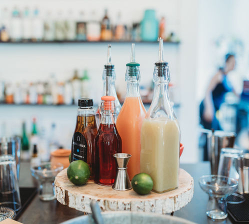 Cocktail station with colorful glass pour bottles of mixers and syrups, two limes and a metal jigger on a rustic wood slice, blurred bar and bartender in the background.