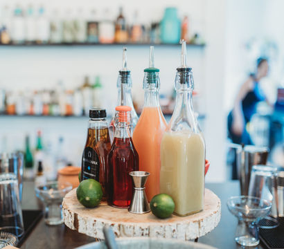 Cocktail station with colorful glass pour bottles of mixers and syrups, two limes and a metal jigger on a rustic wood slice, blurred bar and bartender in the background.