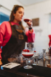 Smoky raspberry coupe cocktail garnished with skewered raspberries on a bar counter, with a blurred bartender in a pink sweatshirt and apron tasting a drink in the background.