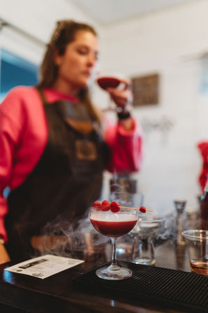 Smoky raspberry coupe cocktail garnished with skewered raspberries on a bar counter, with a blurred bartender in a pink sweatshirt and apron tasting a drink in the background.