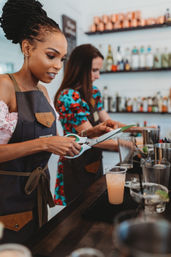 Two bartenders in aprons trimming a green garnish with scissors over a pink cocktail at a craft cocktail bar with bottle-lined shelves