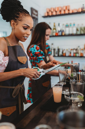 Two bartenders in aprons trimming a green garnish with scissors over a pink cocktail at a craft cocktail bar with bottle-lined shelves