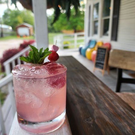 Pink raspberry cocktail with mint garnish on a wooden porch table, outdoor summer scene with blurred colorful chairs and chalkboard sign in the background.