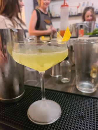 Zesty green-yellow craft cocktail in a coupe glass with a lemon-twist garnish on a bar mat, blurred bartending tools and people in the background