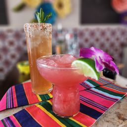 Vibrant pink frozen margarita with lime wedge and purple orchid next to a tall salted‑rim spicy cocktail topped with mint, served on a colorful striped table runner.