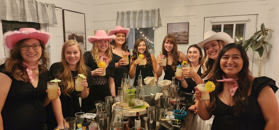 Nine women in cowgirl hats and pink bandanas smiling and raising tropical cocktails around a rustic farmhouse-style bar counter filled with shakers, bottles and garnishes under string lights