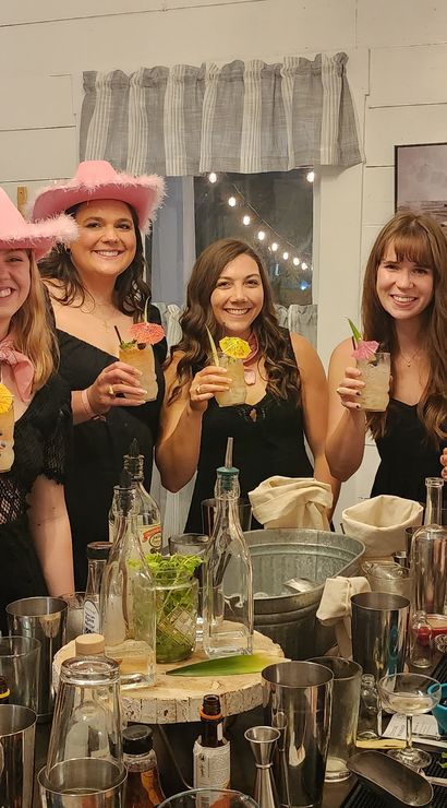 Group of women in black dresses and pink cowgirl hats smiling and holding tropical cocktails around a rustic white farmhouse-style bar with string lights and shiplap walls