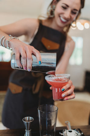 Smiling bartender pours a pink, salt-rimmed cocktail from a metal shaker into a coupe glass at a bar, with colorful nail polish and cocktail tools visible