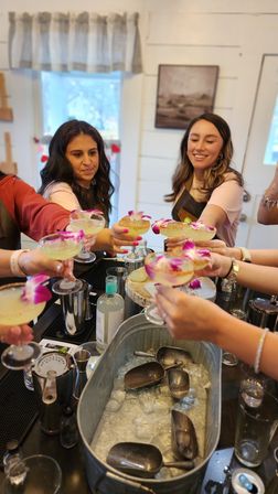 Cheerful group toasting margarita-style cocktails topped with purple orchid garnishes at an indoor cocktail-making party over a bar counter with an ice tub and bartending tools.