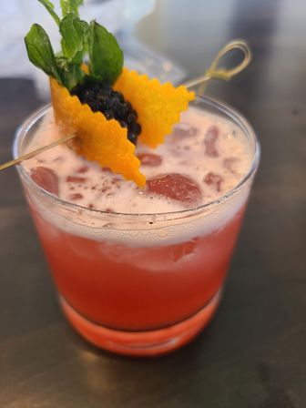 Icy pink cocktail in a short rocks glass with frothy top, garnished with a zigzag orange peel, blackberries and fresh mint on a cocktail pick.
