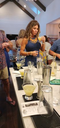 Woman in a navy dress sipping a yellow cocktail at a marble home bar with shiplap walls, exposed wooden beams, cocktail shakers and glasses during a lively gathering.