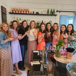 Nine women toasting with pink cocktails at a cozy indoor cocktail workshop bar, smiling in front of shelves of spirits, copper mugs and fresh herbs on the counter