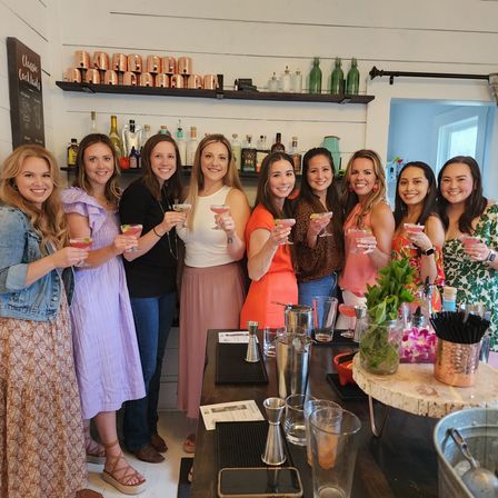 Nine women toasting with pink cocktails at a cozy indoor cocktail workshop bar, smiling in front of shelves of spirits, copper mugs and fresh herbs on the counter
